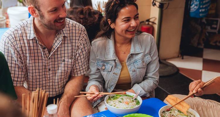 Los viajeros se sientan en banquetas bajas disfrutando de tazones de pho en un pequeño comedor local