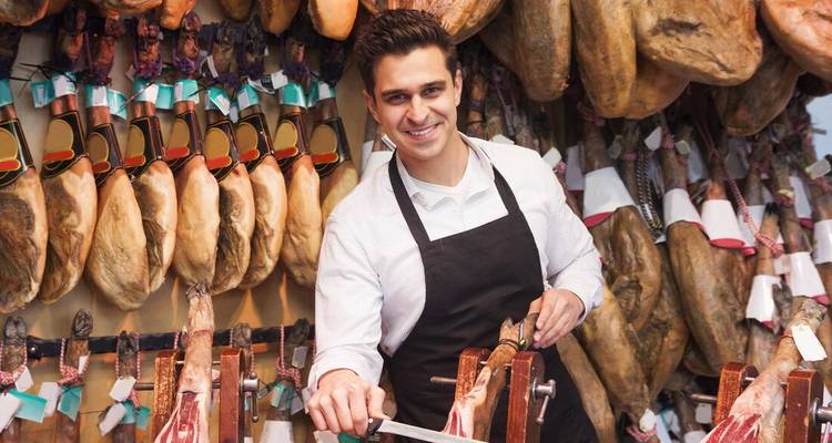 Tendero cortando jamón español rodeado de jamones colgantes.