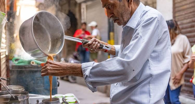 Un vendeur ambulant verse avec dextérité du thé chai chaud d'une casserole dans de petites tasses le long d'une ruelle animée.
