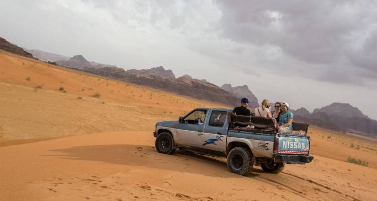 Una camioneta que lleva turistas atraviesa dunas de arena naranja respaldadas por montañas dentadas.