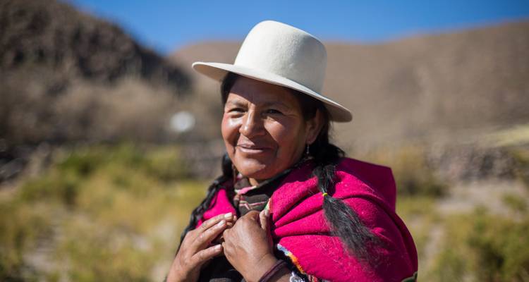 Portrait chaleureux d'une femme andine vêtue d'habits traditionnels et d'un chapeau blanc, souriante devant un paysage montagneux.