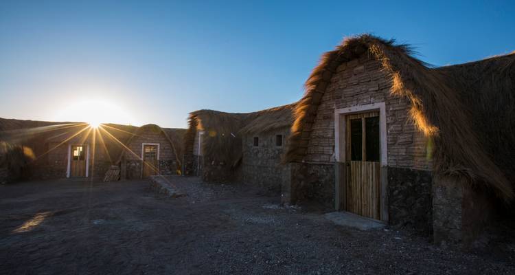 De simples lodges en pierre et chaume brillent sous la faible lumière du lever de soleil sur une plaine bolivienne isolée.