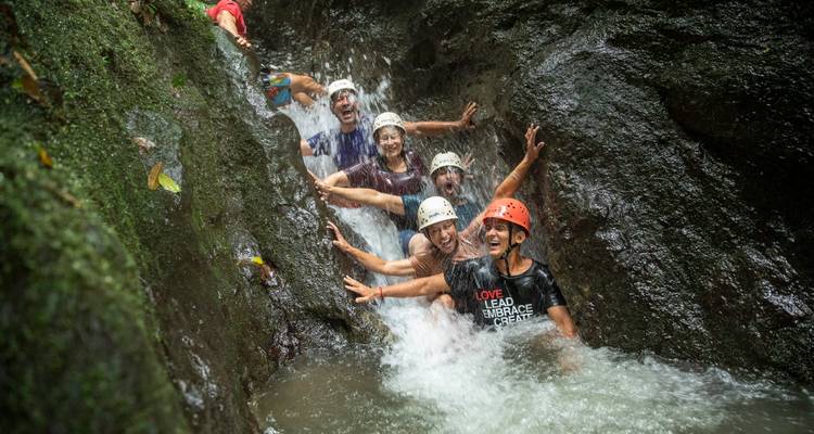 Un groupe portant des casques rit ensemble assis dans un canyon avec cascade tumultueuse lors d'une aventure.