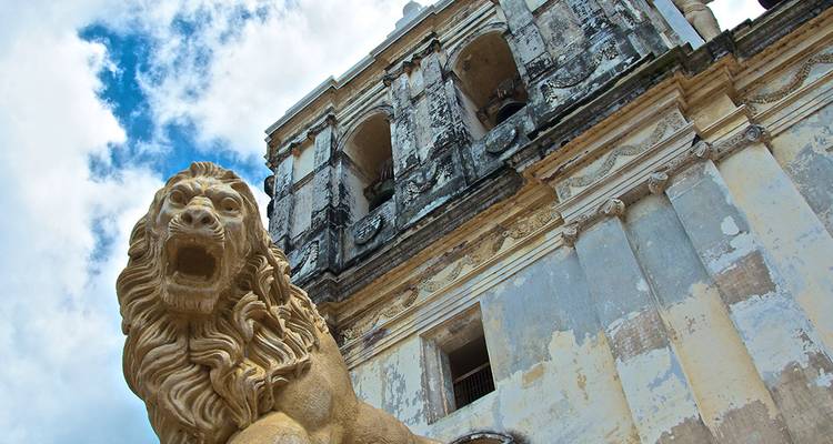 Una estatua de león de piedra que ruge custodia la fachada desgastada por el tiempo de una gran catedral colonial bajo un cielo brillante.