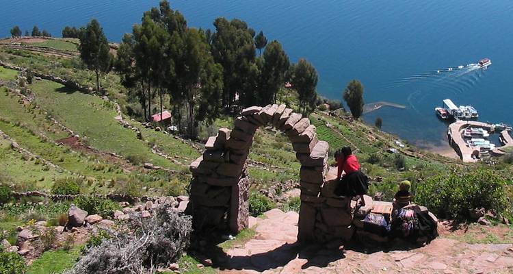 Porche en pierre sur l'île de Taquile encadrant des collines en terrasses et le lac Titicaca scintillant avec des habitants vêtus de costumes traditionnels.