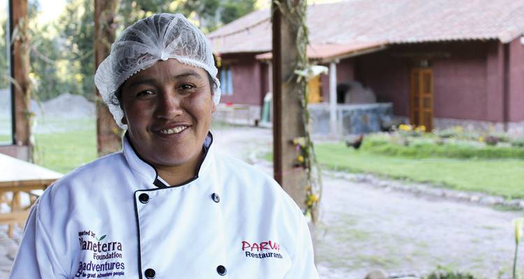 Chef femme souriante vêtue d'une veste blanche et d'un filet à cheveux se tient dans un cadre de restaurant en plein air en milieu rural.