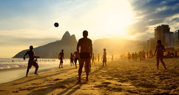 Silhouetten, die bei Sonnenuntergang am Strand Ball spielen, mit Stadtgebäuden