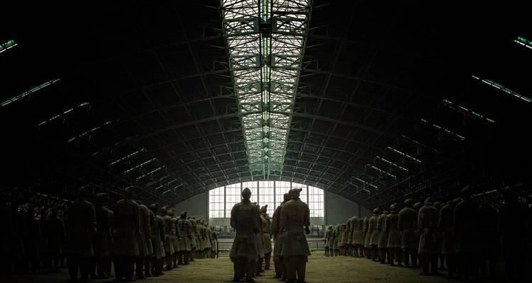 Filas de guerreros de terracota se mantienen en pie bajo una luz baja y dramática debajo de un techo de hangar arqueado y gigantesco