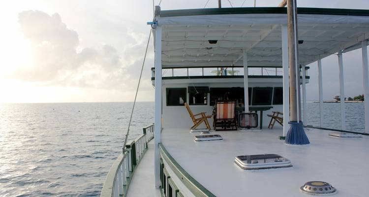 Zijdek van een klein cruiseschip bij zonsopgang met opvouwbare stoelen en zacht licht over een kalme zee.