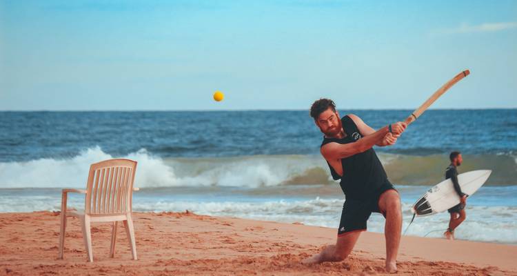 Een man die cricket speelt op het strand met een surfer op de achtergrond.