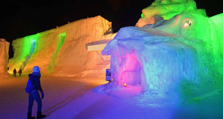 Un visiteur vêtu de vêtements d'hiver explore des grottes de glace brillamment éclairées avec une illumination d'un vert et d'un rose éclatants.