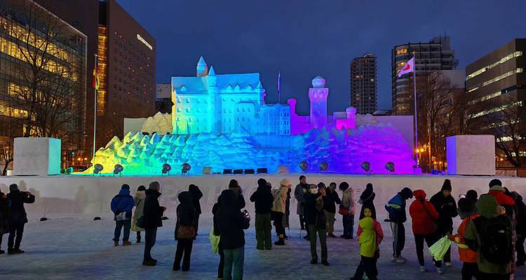 Une foule se rassemble devant une grande sculpture de château de neige illuminée au crépuscule à Sapporo.
