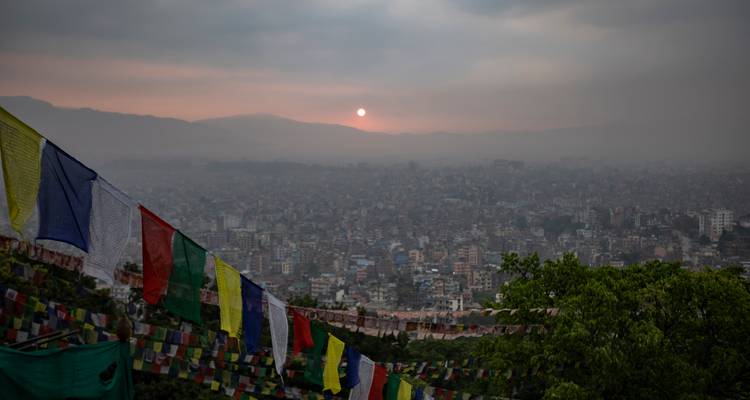 La ciudad de Katmandú se extiende bajo un atardecer brumoso con banderas de oración ondeando en primer plano