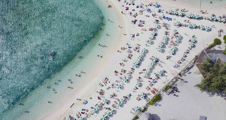 Vista aérea de una playa de arena blanca en forma de media luna con agua turquesa, sombrillas y bañistas.