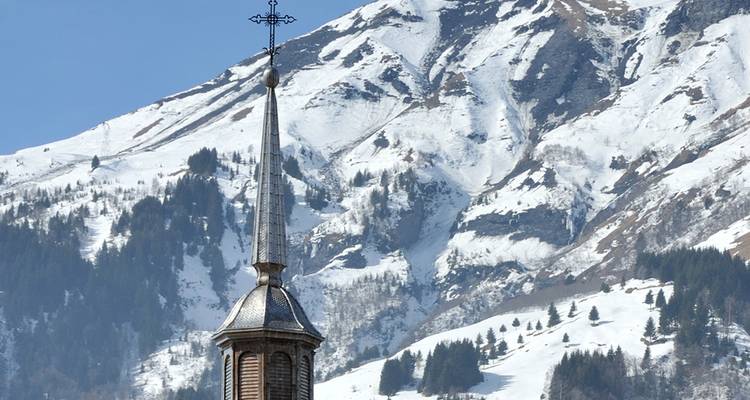 Le haut clocher d'église surmonté d'une croix se dresse devant des pentes alpines enneigées.