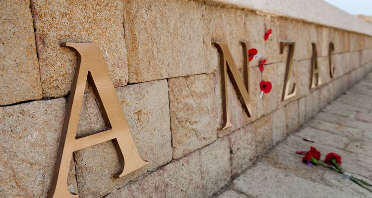 Bronze-ANZAC-Gedenkbuchstaben, die in eine Sandsteinmauer eingelassen sind, mit roten Mohnblumen darunter.