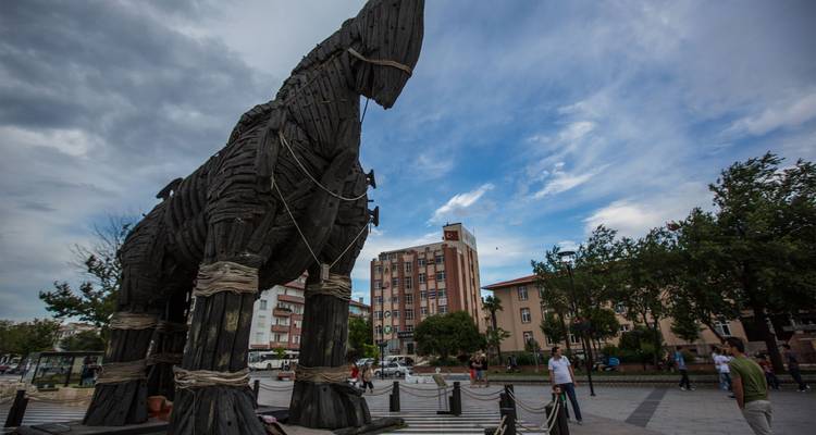 Réplica grande de caballo de Troya de madera exhibida en una plaza del pueblo con gente caminando cerca bajo un cielo parcialmente nublado.