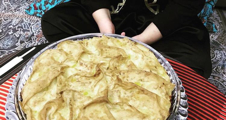 Una bandeja grande de pastel turco horneado descansando en las piernas cruzadas de una mujer sobre una alfombra con diseños.