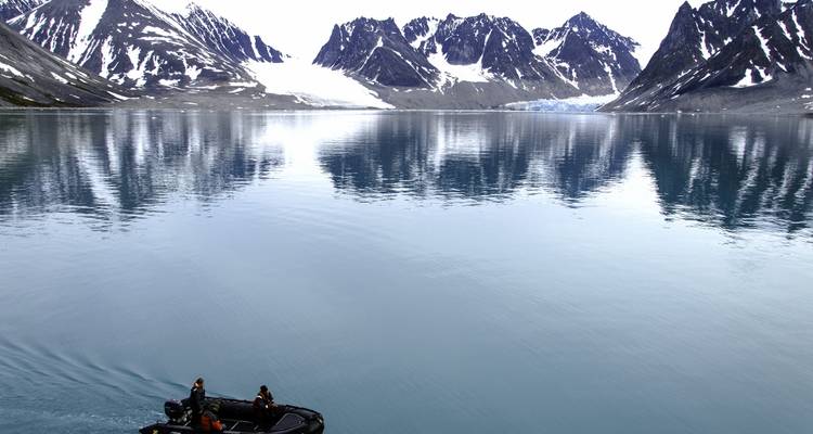 Un petit zodiac avec des touristes navigue à travers un fjord lisse comme un miroir avec des pics enneigés déchiquetés reflétés dans l'eau.
