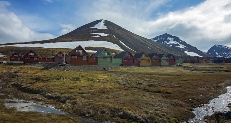 Des rangées de maisons en bois colorées bordent une vallée sous une montagne striée de neige par une journée lumineuse.