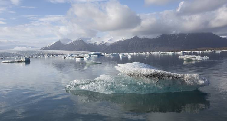 Un iceberg aux reflets bleus flotte dans un lagon glaciaire calme avec des montagnes enneigées et des nuages au loin.