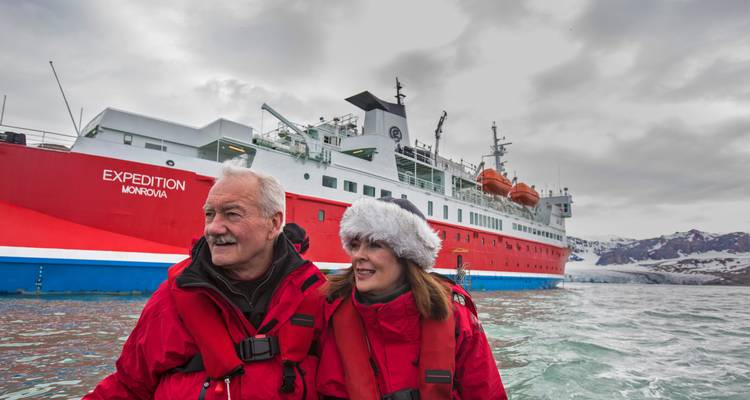 Couple âgé souriant en parkas rouges à bord d'un petit bateau avec un navire d'expédition rouge derrière eux.