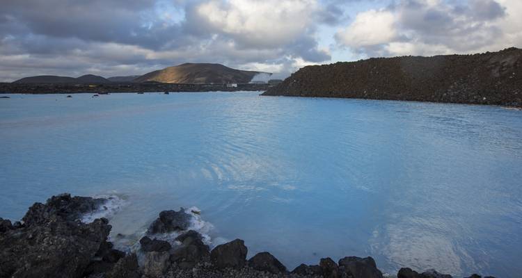 Lagon géothermique bleu laiteux bordé de roches volcaniques sombres sous un ciel maussade.