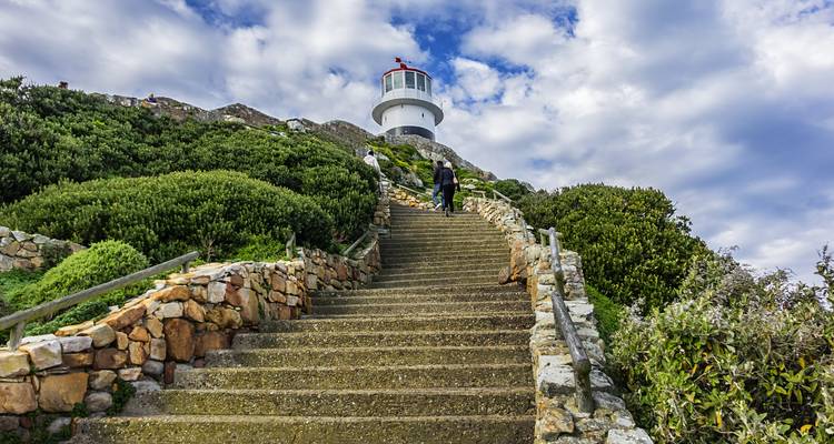 Long escalier en pierre menant à un phare blanc sur un promontoire rocheux de Cape Point entouré de buissons verts et de nuages spectaculaires.