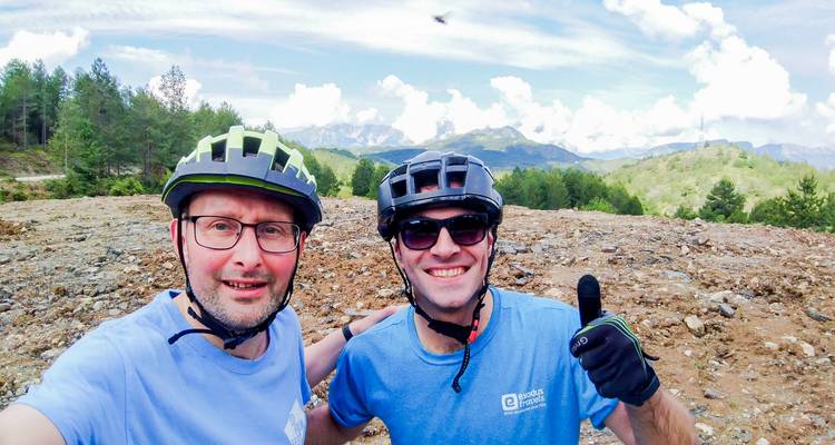 Twee wielrenners die een vrolijk selfie maken op rotsachtig terrein met op de achtergrond de bergen van Albanië.