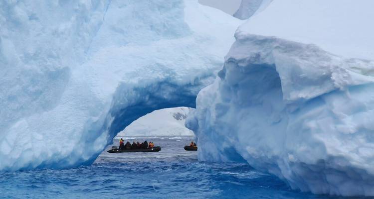 Zodiac boat of explorers passing beneath towering blue ice arch on rough sea