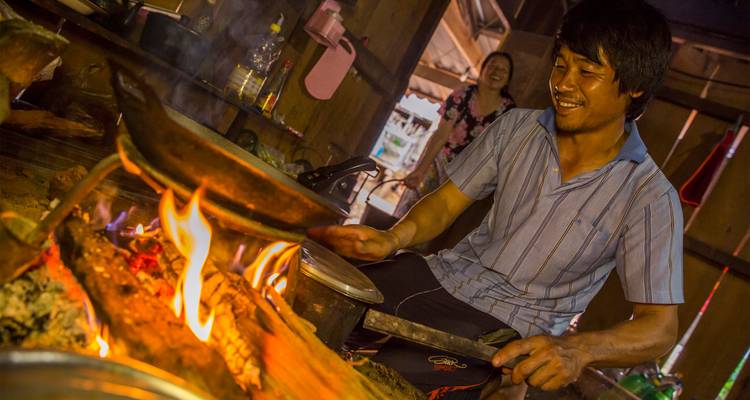 Le cuisinier local sourit en remuant les aliments sur un feu ouvert à l'intérieur de la cuisine traditionnelle du village en bois.