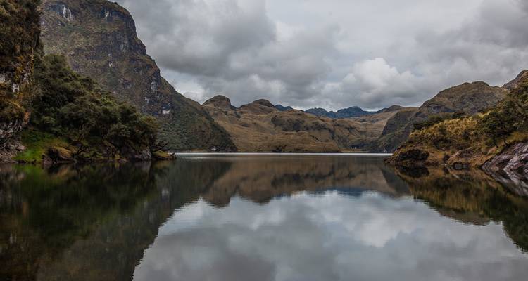 Stiller Bergsee, der einen bewölkten Himmel und raue Moorlandhänge widerspiegelt, umgeben von hohen Andengipfeln.