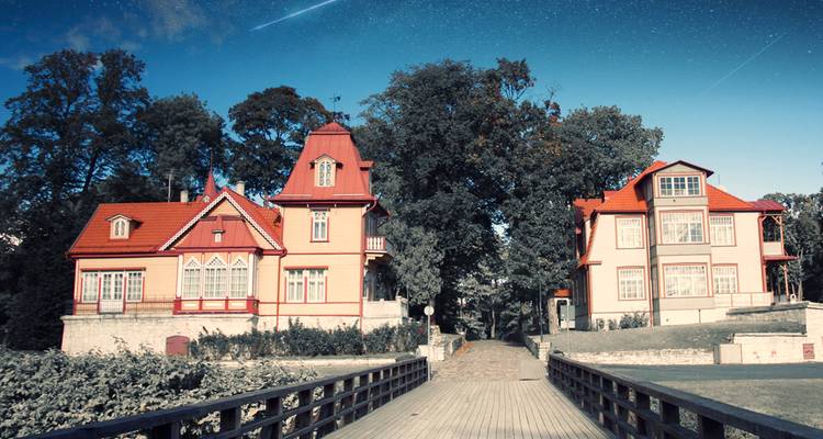 De pittoresques villas en bois aux toits rouges se dressent à l'extrémité d'une passerelle en bois entourées d'arbres feuillus sous un ciel bleu dégagé.