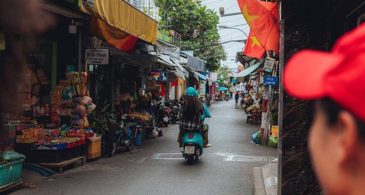 Een scooter rijdt door een smalle straat vol winkels en Vietnamese vlaggen.