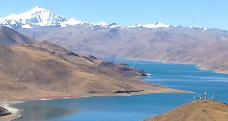 Hochgelegener See mit schneebedeckten Himalaya-Gipfeln jenseits von sanften braunen Hügeln unter klarem Himmel.