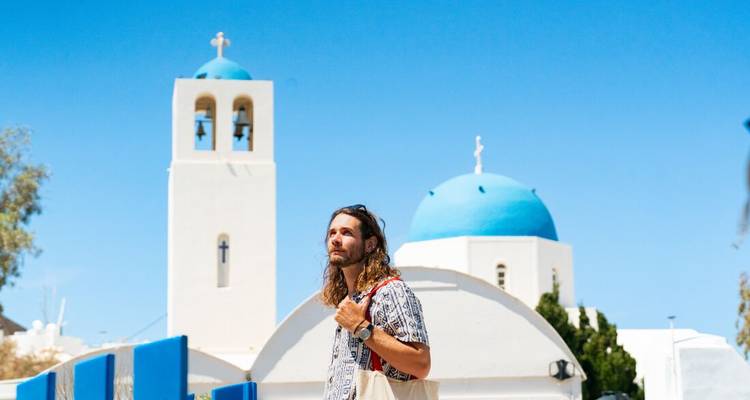 Reisender vor einer weißen Kirche mit blauen Kuppeln und Glockenturm unter lebendigem Himmel