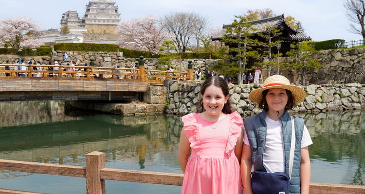 Dos niños están de pie junto al foso con flores de cerezo enmarcando el icónico Castillo Blanco de Himeji detrás de ellos.