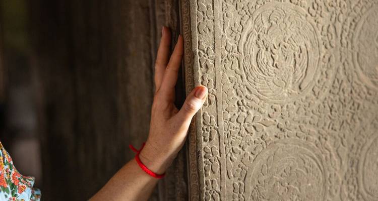 Una mano con una pulsera roja toca suavemente intrincados tallados de piedra en una pared de templo jemer antiguo.