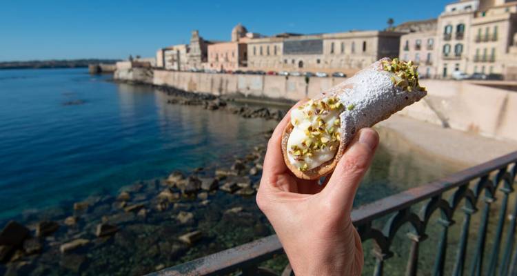 Una mano sostiene un cannolo cubierto de pistachos con el frente de agua de Ortigia y sus edificios históricos de fondo.