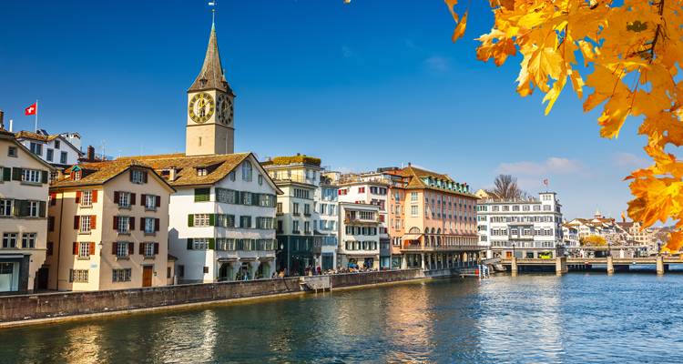 Kleurrijke huizen aan de rivier en de kerktoren van St. Peter ingekaderd door gouden herfstbladeren in Zürich.