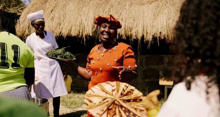 Femme locale en robe orange présentant fièrement des légumes verts frais lors d'une démonstration culinaire culturelle dans un village au toit de chaume