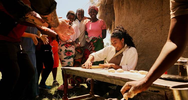 Touristes et femmes locales souriantes en train de préparer de la pâte en plein air à côté d'une hutte au toit de chaume sous un soleil éclatant