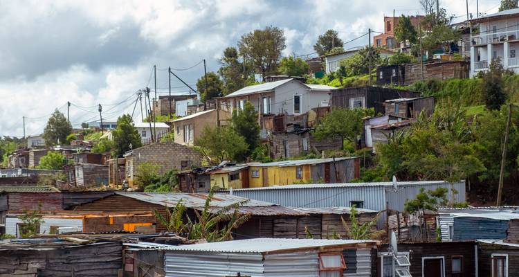 Établissement informel sur une colline composé de maisons en tôles ondulées et briques avec des lignes électriques aériennes sous un ciel nuageux menaçant.