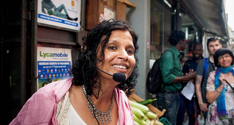 Un guide touristique souriant portant un casque-micro s'adresse aux visiteurs dans un marché de rue étroit et animé.
