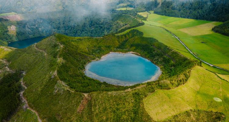 Vue aérienne d'un lac de cratère volcanique circulaire entouré de collines verdoyantes et de nuages brumeux sur São Miguel.
