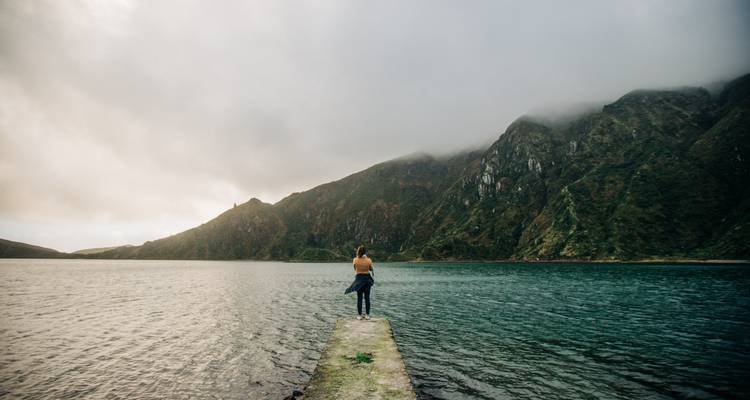 Un voyageur solitaire se tient sur une jetée étroite en béton qui s'avance dans un lac de montagne brumeux sous des nuages bas.