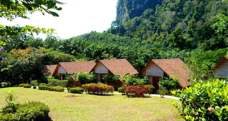 Fila de pequeños bungalows de madera con techos rojos rodeados de jardines cuidados y con acantilados boscosos empinados de fondo.