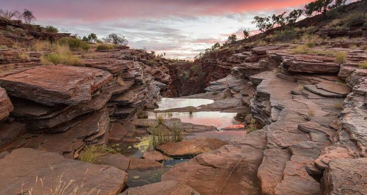 Un desfiladero rojo escarpado con pozas rocosas resplandece bajo cielos de amanecer pastel en el Parque Nacional Karijini.