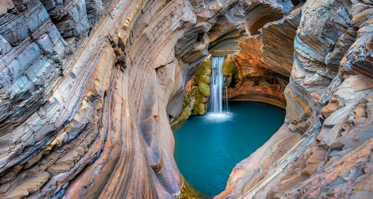Una piscina natural turquesa alimentada por una cascada delicada se encuentra dentro de paredes rocosas con bandas en espiral.