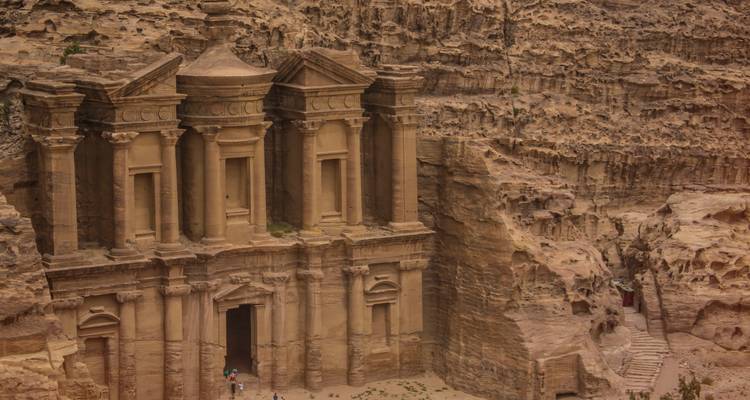 La gran fachada de piedra tallada del Monasterio de Petra se alza desde acantilados desérticos accidentados mientras unos pocos turistas están de pie en la entrada de abajo.